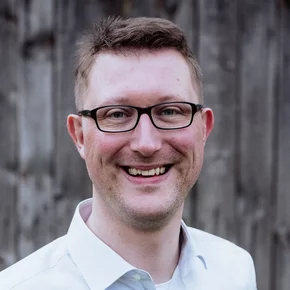 Ein lächelnder Mann mit Brille trägt ein helles Hemd. Er steht vor einer holzverkleideten Wand. Sein freundlicher Gesichtsausdruck vermittelt eine positive Stimmung.