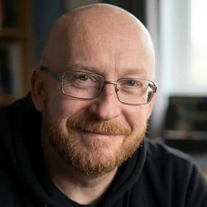 Ein lächelnder Mann mit Glatze und Brille sitzt an einem Schreibtisch. Er trägt einen schwarzen Hoodie. Im Hintergrund sind unscharfe Computerbildschirme und Buchregale sichtbar. Das Bild vermittelt eine freundliche, einladende Atmosphäre.