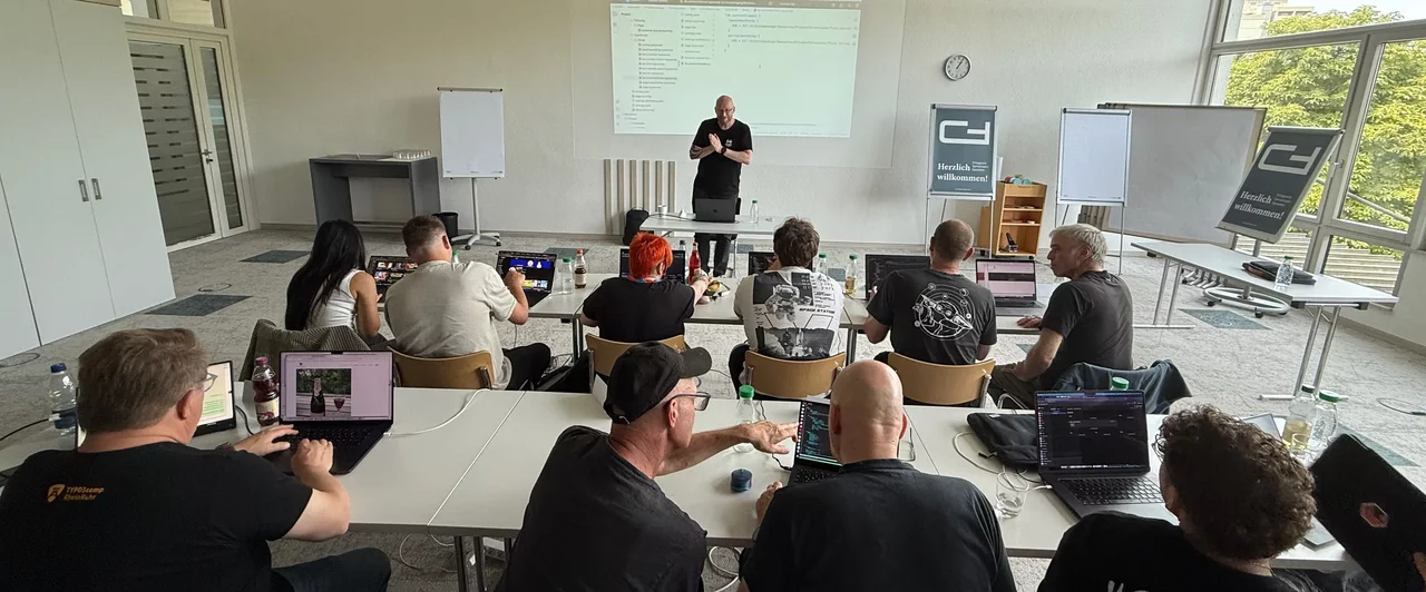 Eine Gruppe von Personen sitzt in einem Seminarraum und hört Wolfgang Wagner zu, der vorne an einem Tisch steht. An der Wand hinter ihm ist eine Präsentation zu sehen. Mehrere Teilnehmer arbeiten an Laptops. Tageslicht strömt durch große Fenster.